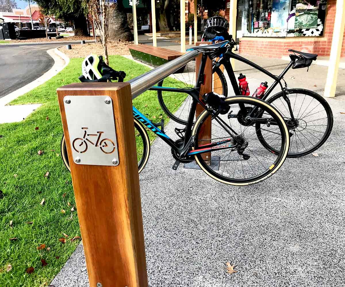 Two bicycles are secured to a bike rack in an urban setting with storefronts nearby.