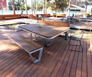 Outdoor seating area in a school with wooden tables and benches on a deck, surrounded by trees.