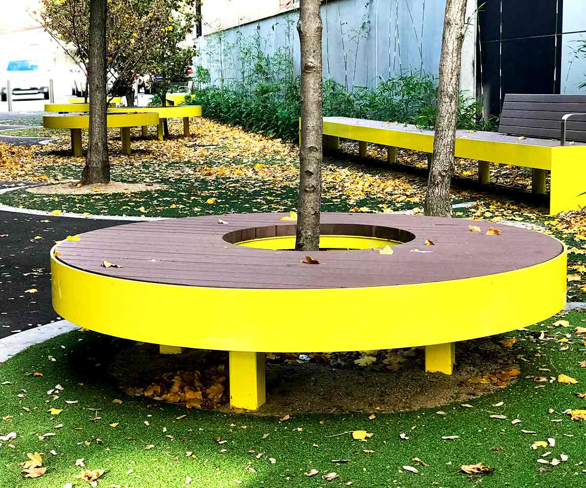 A modern park scene with circular tree benches and a long bench, all with wooden slats and a yellow frame.