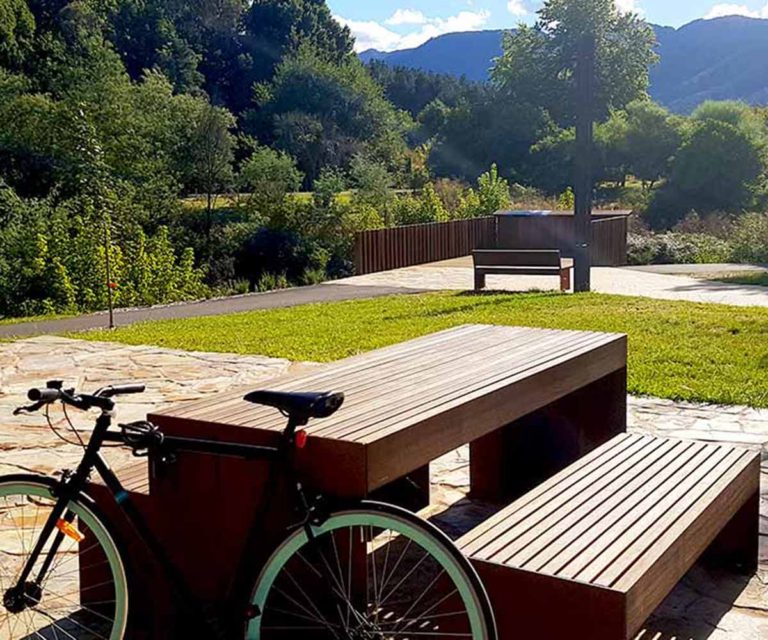 Timber outdoor picnic table with bench seating in a park, with a bicycle nearby and mountains in the background.