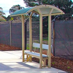 An outdoor park shelter with a metal bench and a curved roof, surrounded by a fence and greenery