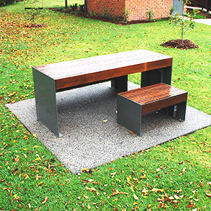 A modern outdoor picnic table with a bench on a stone slab in a grassy park. The table has a minimalist wood and metal design.