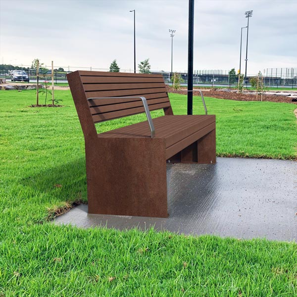 A brown metal bench with engraved paw prints sits on a concrete path in a green dog park, under a cloudy sky.