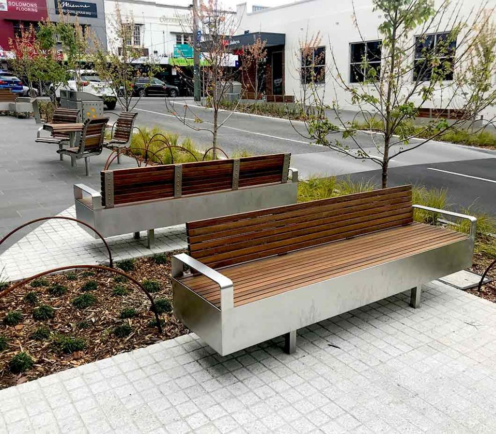 Public benches with wooden slats and metal frames and armrests in a streetscape.
