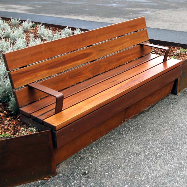 A wooden park seat with armrests placed on a gravel path in front of a landscaped area with plants.