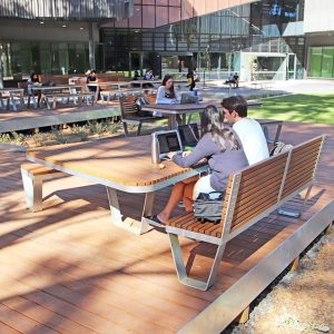 Monash University Outdoor seating area with wooden benches and tables on a terrace. People working on laptops and studying.