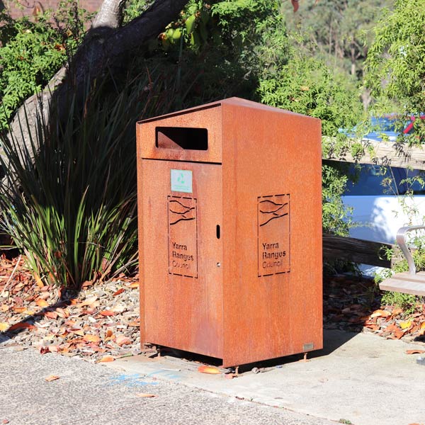 A rust-colored metal bin labelled "Yarra Ranges Council" in a public area, surrounded by greenery and a footpath.
