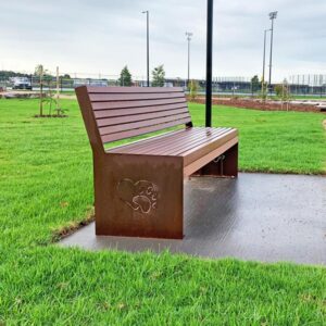 A brown metal bench with engraved paw prints sits on a concrete path in a green dog park, under a cloudy sky.