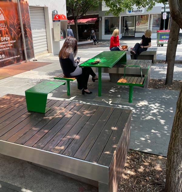 People sitting at green metal and wooden outdoor tables in a shaded urban area. Trees and storefronts are visible in the background.