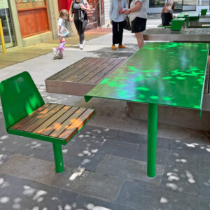 A green outdoor bench with wooden slats is attached to a table in a busy street. People are walking and using street furniture in the background.