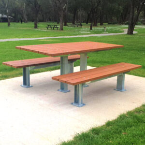 A wooden outdoor picnic table with attached benches on a concrete slab, surrounded by green grass and trees.