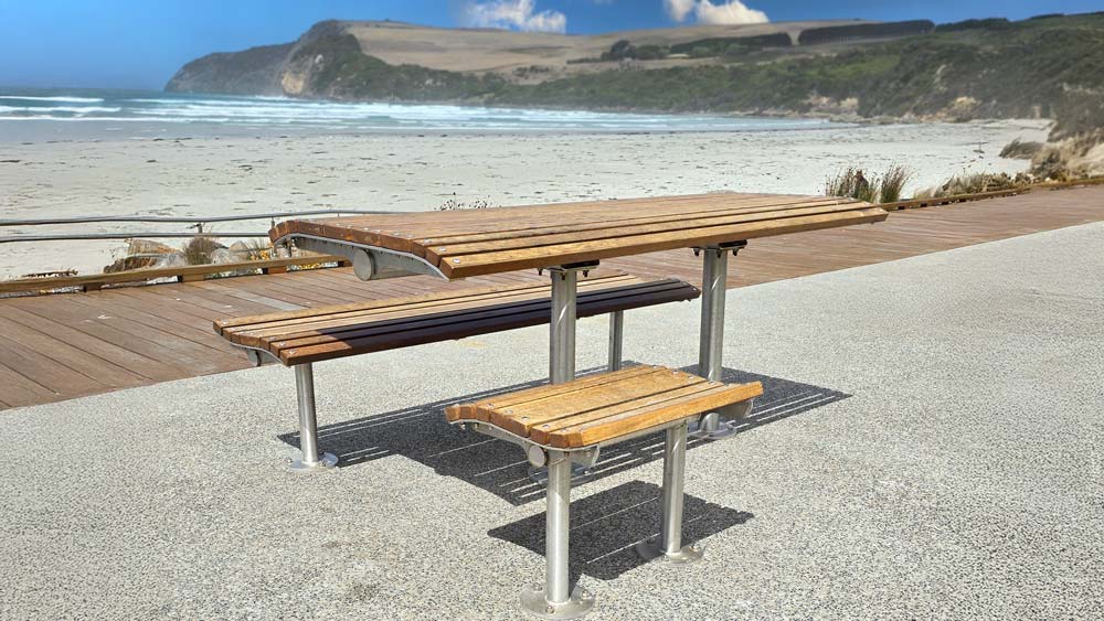 Wooden picnic table with metal legs on a paved seaside path. Picnic table installed by Draffin Street Furniture.