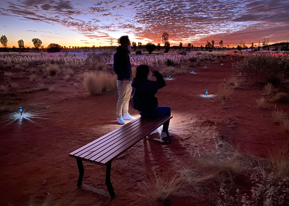 Two people gaze at a sunset over the Field of Light installation in Uluru. One person is sitting on a bench.