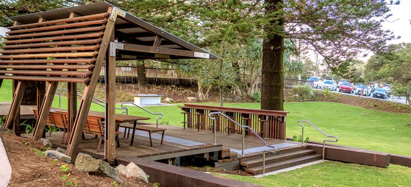 A shade structure with a slatted roof and benches on a deck in a lush park.