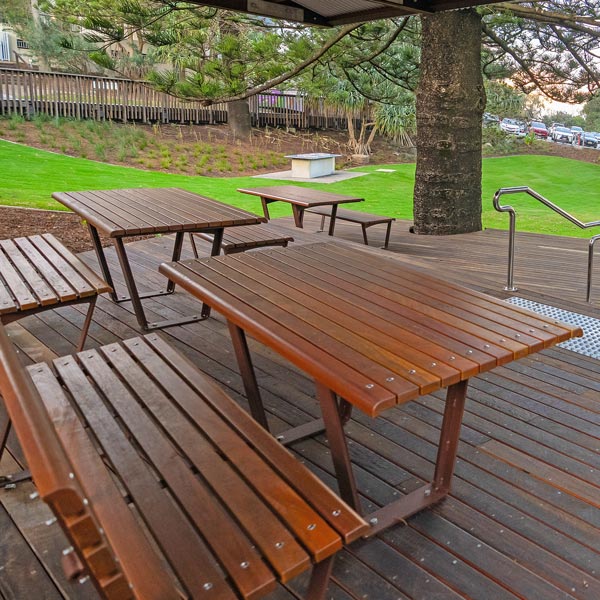 Wooden park benches and picnic tables on a deck surrounded by green grass and tall trees.