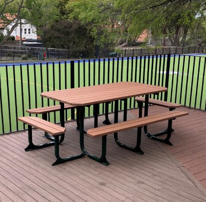 A picnic table with attached benches on a wooden deck. The benches have 3-leg supports. A sports field with trees in the background.
