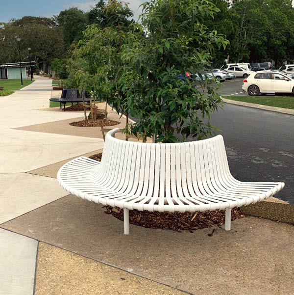 Curved white tree seat encircling a tree on a sidewalk, with parked cars and greenery in the background.