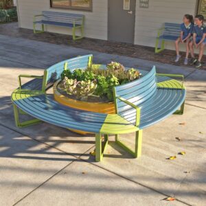 Modular curved seats and benches arranged to form a circular shape surrounding a large planter with herbs. School ground office in the background.